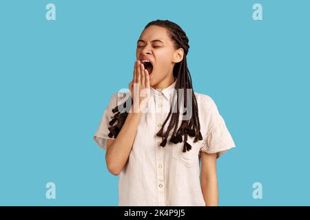 Junge erschöpfte Frau mit schwarzen Dreadlocks, die den Mund mit der Hand bedeckt, hält die Augen geschlossen, fühlt sich müde oder müde, trägt weißes Hemd. Innenaufnahme des Studios isoliert auf blauem Hintergrund. Stockfoto