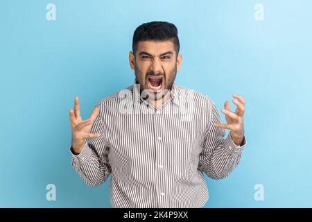 Wütender, gutaussehender Geschäftsmann, der die Kamera anschaut, einen aggressiven Gesichtsausdruck hat, negativ, Hass ausdrückt und gestreiftes Hemd trägt. Innenaufnahme des Studios isoliert auf blauem Hintergrund. Stockfoto