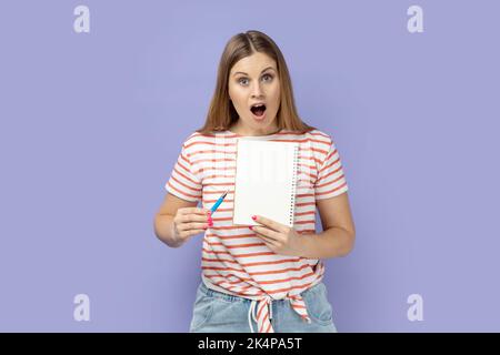 Porträt der erstaunt überrascht blonde Frau trägt gestreiftes T-Shirt mit Organizer mit leerem Papier mit Kopieplatz für Werbung. Innenaufnahme des Studios isoliert auf violettem Hintergrund. Stockfoto