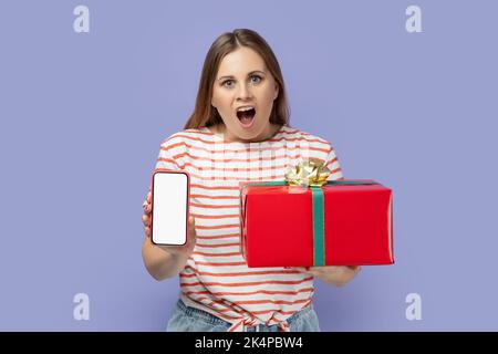 Erstaunt aufgeregt Frau trägt gestreiftes T-Shirt mit roten Geschenk-Box und zeigt Handy mit weißem Bildschirm mit Kopieplatz für Werbung. Innenaufnahme des Studios isoliert auf violettem Hintergrund. Stockfoto