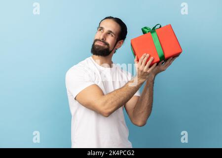 Porträt eines neugierigen Mannes mit Bart trägt weißes T-Shirt schütteln rote Geschenkbox mit grünem Band, versuchen zu erraten, was drin ist, Überraschung. Innenaufnahme des Studios isoliert auf blauem Hintergrund. Stockfoto