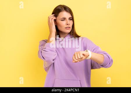 Porträt einer hübschen Frau, die auf die Armbanduhr schaut, die einen Termin vereinbart, sich trifft, ihr Haar mit der Hand berührt und einen violetten Hoodie trägt. Innenaufnahme des Studios isoliert auf gelbem Hintergrund. Stockfoto