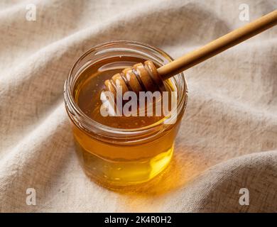 Honig im Glasbehälter und hölzerne Honigtaube auf Leinenhintergrund. Honig tropft aus dem Honigtauer. Stockfoto