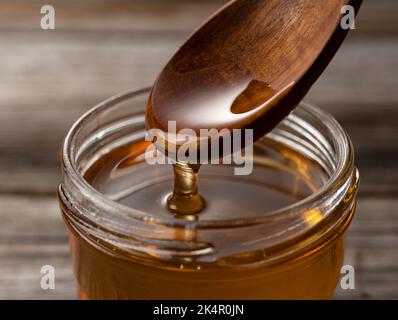 Honig- und Holzlöffel im Glasbehälter auf altem Holzhintergrund. Honig tropft vom Löffel. Stockfoto