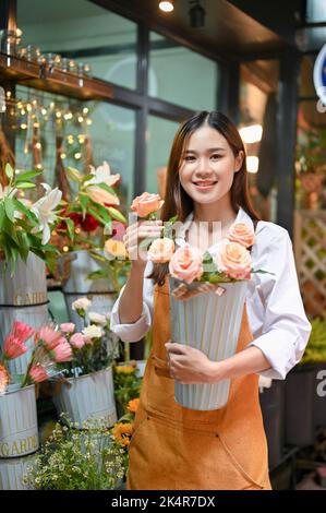 Porträt, schöne attraktive junge asiatische Floristin oder Blumenhändler mit einer schönen Rosenvase, die in ihrem Blumenladen steht. Stockfoto