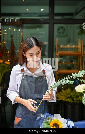 Portrait, attraktive junge asiatische Floristin, die mit einer Schere einen Strauß macht und in ihrem Laden Blütenstiele schneidet. Kleinunternehmer Konzept Stockfoto
