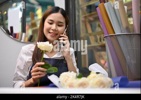 Schöne junge asiatische weibliche Floristin oder Blumenhändler sprechen am Telefon, nehmen Bestellung von ihrem Kunden während der Arbeit in ihrem Blumenladen. Stockfoto