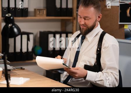 Detective, der am Schreibtisch in seinem Büro mit Dokumenten arbeitet Stockfoto