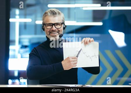 Ein Mann mit Kopfhörern und Mikrofon. Im Büro sitzen, Grafiken halten, ein Projekt in den Händen halten und mit einem Stift auf die Kamera zeigen. Führt Geschäftstreffen, Webkonferenzen und lächelt. Stockfoto