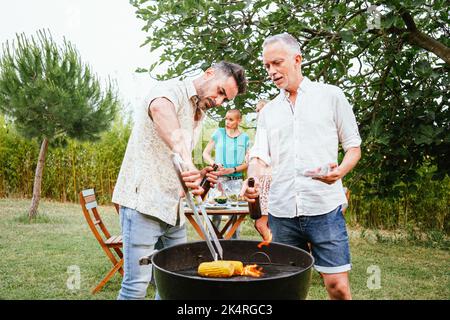 Zwei Männer, die auf dem Land beim Grillen Bier kochen und toasten. Im Hintergrund sprechen drei Freundinnen und haben Spaß. Lifestyle-Konzept. Stockfoto