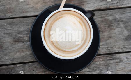 Tasse Cappuccino aus Latte auf einem Vintage-Holztisch in Nahaufnahme. Üppiger Milchschaum sprudelt. Köstlicher Morgendrink Stockfoto