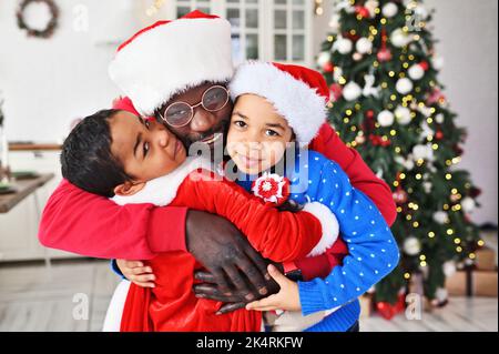 Vater und Kinder - eine Tochter und ein Sohn in einem Weihnachtsmann-Kostüm mit einem gefälschten Bart, der vor dem Hintergrund des Weihnachtsbaums umarmt und lächelt. Stockfoto