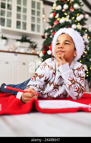 Der Junge im weihnachtsmütze, der auf einem karierten Tuch liegt, lächelt träumerisch und schreibt einen Brief an den Weihnachtsmann auf ein weißes Blatt - eine Wunschliste auf dem Hintergrund eines Weihnachtsfestes Stockfoto