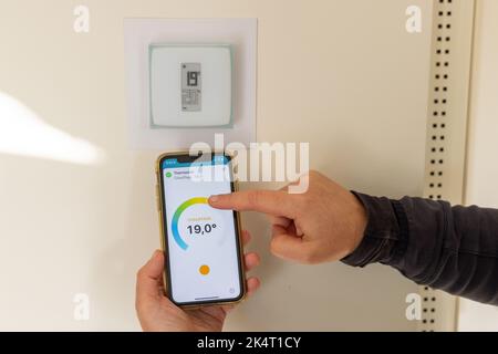Person, die ein Smartphone in der Hand hält, um ihren angeschlossenen Thermostat in einem französischen Haus einzustellen Stockfoto