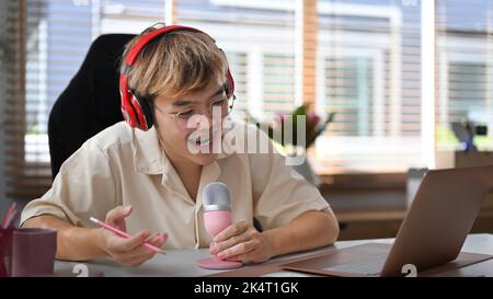 Fröhlicher junger asiatischer Mann mit Laptop und Mikrofon Streaming Audio-Podcast zu Hause Studio Stockfoto