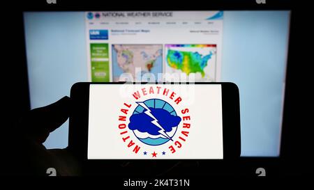 Person, die das Mobiltelefon mit dem Logo der amerikanischen Agentur National Weather Service (NWS) auf dem Bildschirm vor der Webseite hält. Konzentrieren Sie sich auf die Telefonanzeige. Stockfoto