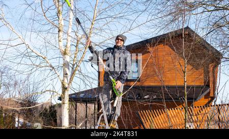 Ein Gärtner steht auf einer Stehleiter mit einer elektrischen Kette, einem Gartenscherer mit Teleskopstange und Schneidezweigen in Winterausrüstung. Wint Stockfoto