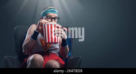 Lustiger Superheld, der Horrorfilme ansieht und Popcorn isst, ist erschrocken und versteckt sich Stockfoto