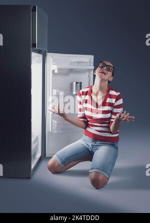 Verzweifelte weinende Frau und leerer Kühlschrank, sie ist hungrig, aber sie hat zu Hause kein Essen Stockfoto