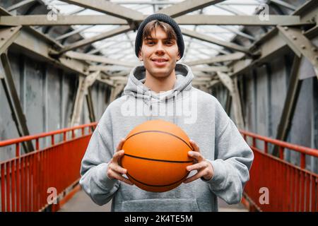 Junger sportlicher Mann hält Basketballball auf städtischem Hintergrund. Konzept des aktiven Lebensstils. Stockfoto