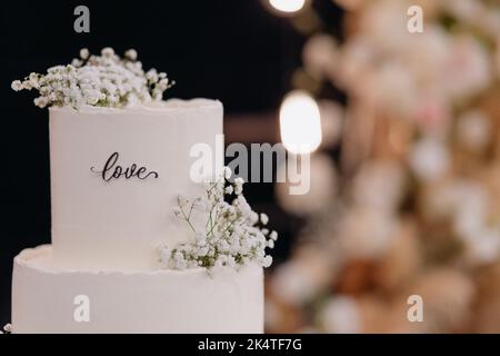 Nahaufnahme einer weißen Hochzeitstorte, die mit Blumen geschmückt ist, die draußen auf einem Stand mit Geschirr vor dem Hintergrund eines Abendbogens stehen. Stockfoto