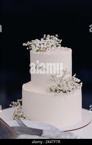 Am Abend steht auf einem Stand mit Utensilien ein mit Blumen geschmückter weißer Hochzeitstorte aus nächster Nähe. Stockfoto