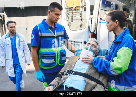 Arzt, der mit Atemschutzmaske auf der Krankenwagen-Trage zum Seniorenpatienten eilte, der von Sanitätern im Krankenwagen gebracht wurde. EMS Stockfoto