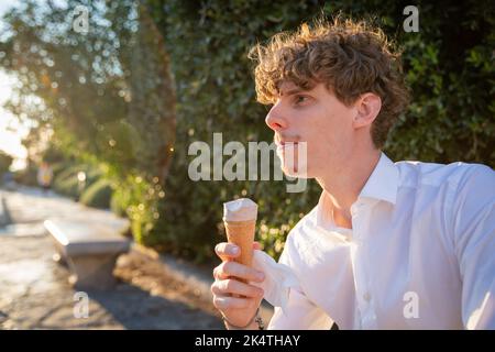 Junger Mann, der an einem sonnigen Tag auf einer Bank in einem öffentlichen Park sitzt, genießt Eis, Kopierplatz links. Stockfoto