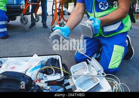 Rettungsassistent hält Defibrillator-Pads in den Händen, während der Rettungsdienst im Freien in der Nähe des Rettungswagens verletzte. Erste Hilfe durchführen Stockfoto