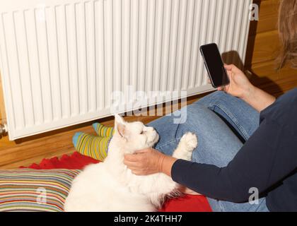 Frau und ihre weiße Katze sitzen in der Nähe der Zentralheizungsbatterie. Sie wärmt seine Füße in gestreiften Socken auf der Batterie. Sanktionen, Russland. Konzept Wintersaison Stockfoto