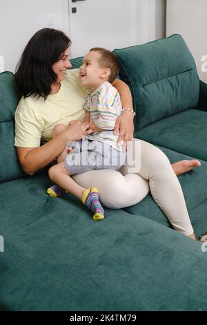 Mutter hält Kind mit Behinderung auf dem Sofa leben Frau umarmt Jungen mit zerebraler Lähmung zu Hause Mutter Spaß haben, kümmert sich um Jungen, die hat Stockfoto