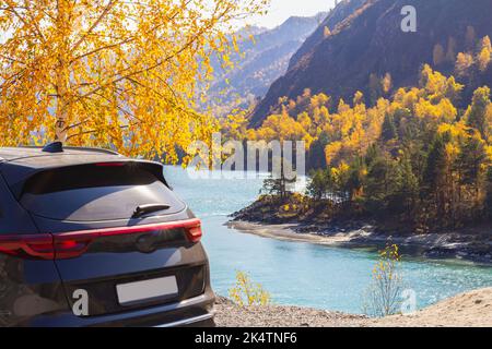 Rückansicht des schwarzen Autos, geparkt in Rastplatz oder Aussichtsplattform, im Schatten der gelben Birkenblätter, des bergblauen Flusses und der gelben Herbsthänge des Berges Stockfoto