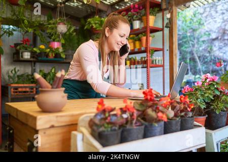Pflanzenverkäuferin mit Handy und Computer im Blumengeschäft, der bestellt Stockfoto