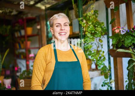 Lächelnde junge Gärtnerin oder Verkäuferin mit grüner Schürze im Blumenladen Stockfoto