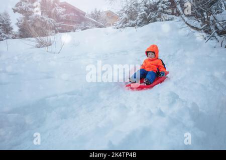 Süßer Junge im orangen Skisport-Outfit geht auf dem Schlitten bergab Stockfoto