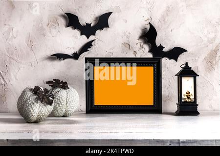 Schwarzer Rahmen mit orangefarbenem Platz für Text, dekorative Kürbisse, Fledermäuse, Laterne auf weißem Holztisch auf lichtstrukturiertem Hintergrund. Halloween-Heimtextilien Stockfoto