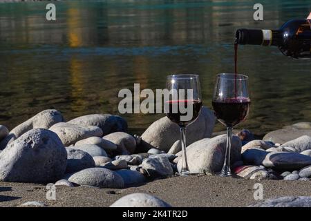 Gießt Rotwein aus der Flasche in Gläser am felsigen Strand des Gebirgsflusses. Zwei Gläser Wein. Romantischer Urlaub oder Weinbereitung Konzept. Natürlicher Rückengr Stockfoto