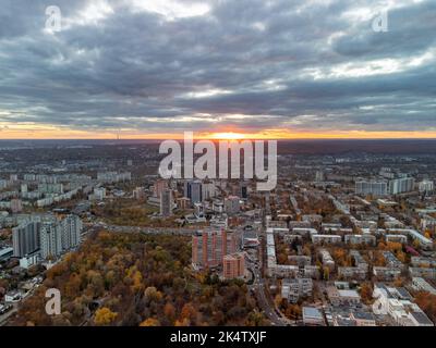Luftaufnahme des herbstlichen Stadtparks mit Blick auf den Sonnenuntergang und epischer Wolkenlandschaft. Botanischer Garten und Kharkiv Stadtzentrum. Wohnviertel Gebäude und Autos fahren Stockfoto