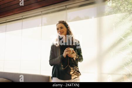 Glückliche ältere Frau lächelt, während sie eine Tasse Tee in ihrem Wohnzimmer hält. Fröhliche ältere Frau, die einen entspannten Morgen zu Hause hat. Stockfoto