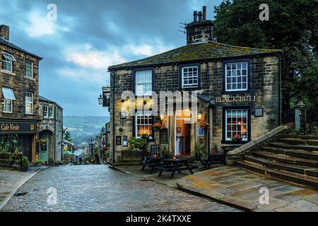 Haworth Main Street (steiler Hügel, alte Gebäude, blaues Abendlicht, historisches Bronte-Schwesterndorf, Pub der Klasse 2) - West Yorkshire, England, Großbritannien. Stockfoto