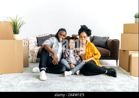 Positive glückliche afroamerikanische Familie zieht in ihr neues Zuhause. Mama Papa und ihre Tochter, sitzen auf einem Boden zwischen Pappkartons im Wohnzimmer, Mädchen zeigt die Schlüssel, sie schauen in die Kamera, lächeln Stockfoto