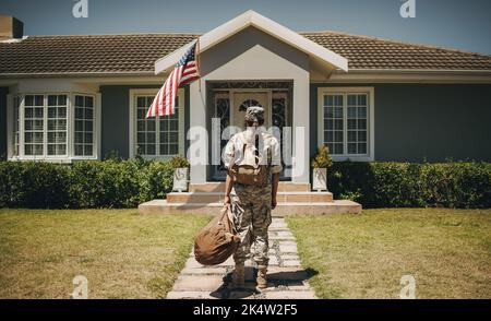 Rückansicht einer patriotischen Soldatinnen, die mit ihrem Gepäck vor ihrem Haus steht. Mutige Militärangehörende, die nach dem Dienst an ihrem Coun nach Hause zurückkehrt Stockfoto