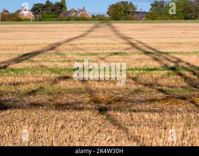 Ich liebe Strommasten; ihre abstrakten, vergilbten Formen finde ich unendlich faszinierend. Hier sehen wir Schatten, die von einem großen Pylon in einem Feld im ländlichen Ra geworfen werden Stockfoto