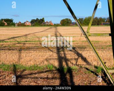 Ich liebe Strommasten; ihre abstrakten, vergilbten Formen finde ich unendlich faszinierend. Hier sehen wir Schatten, die von einem großen Pylon in einem Feld im ländlichen Ra geworfen werden Stockfoto