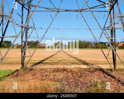 Ich liebe Strommasten; ihre abstrakten, vergilbten Formen finde ich unendlich faszinierend. Hier sehen wir Schatten, die von einem großen Pylon in einem Feld im ländlichen Ra geworfen werden Stockfoto