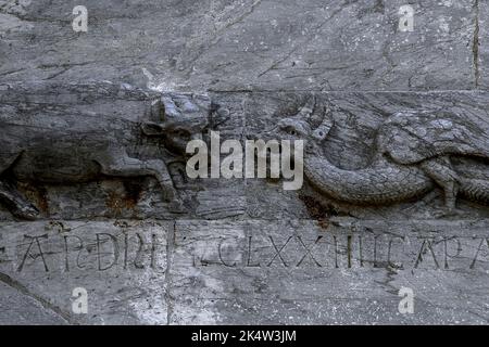 Ein Stier steht vor einem geflügelten, gehörnten Drachen: Detailansicht der romanischen Skulptur aus dem 12.. Jahrhundert in Pisaner Baukunst neben dem Eingang zum Torre Pendente oder schiefen Turm von Pisa, dem geneigten campanile oder Glockenturm auf der Piazza dei Miracoli in Pisa, Toskana, Italien. Die lateinische Inschrift unten dokumentiert die Gründung des Turms im August 1174. Stockfoto