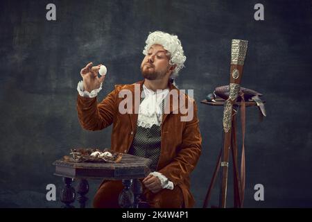 Vintage-Porträt eines jungen Mannes in braunem Vintage-Anzug und weißer Perücke wie mittelalterliche königliche Jäger isoliert auf dunklem Hintergrund. Stockfoto