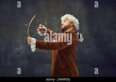 Bogenschießen. Vintage-Porträt eines jungen Mannes in braunem Vintage-Anzug und weißer Perücke wie mittelalterliche königliche Jäger isoliert auf dunklem Hintergrund. Stockfoto