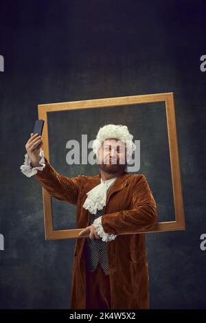 Posieren. Vintage-Porträt eines jungen Mannes in braunem Vintage-Anzug und weißer Perücke wie mittelalterliche königliche Jäger isoliert auf dunklem Hintergrund. Stockfoto