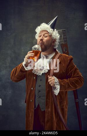 Süße Getränke. Lächelnder junger Mann in braunem Vintage-Anzug und weißer Perücke wie ein mittelalterlicher königlicher Jäger, der Limonade auf dunklem Hintergrund trinkt. Stockfoto